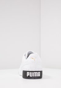Zapatilla blanca con superficie texturizada, suela negra y marca "PUMA". Presenta un acento de logo dorado, diseño redondeado y collar acolchado.