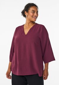 Haut bordeaux à col en V avec des manches trois-quarts, en tissu texturé, coupe décontractée et ourlet arrondi. Porté avec un pantalon noir.