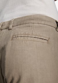 Primer plano del bolsillo trasero de pantalones beige con trabilla para cinturón y camisa blanca metida en la cintura.
