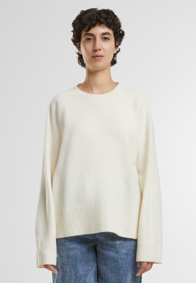 Cream-colored knit sweater with a round neck, oversized fit, ribbed hem, and long sleeves. Soft texture with subtle flecks.