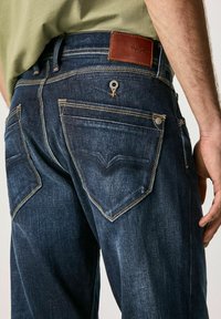 Jeans de mezclilla azul oscuro con costuras claras, que cuentan con dos bolsillos traseros y un parche de cuero marrón en la cinturilla.
