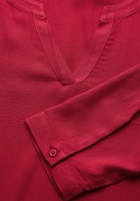 Blusa rossa con scollo a V, caratterizzata da una texture liscia e maniche lunghe. La manica include un dettaglio con bottone vicino al polsino.