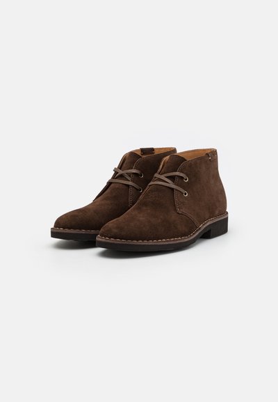 Botines de ante marrón con puntera redonda, que presentan cordones marrones y costuras sutiles, montados sobre un tacón bajo.