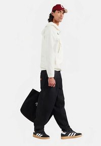 Witte jas met capuchon, zwarte wijde broek, zwarte sneakers met witte strepen, draagt een zwarte draagtas, bordeauxrode pet met logo.