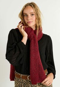Bufanda de punto burdeos con un sutil brillo, de textura suave, caída alrededor del cuello, combinada con una blusa negra texturizada y pantalones de estampado de leopardo.