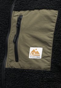 Μαύρο μπουφάν από φλις με υφή, που διαθέτει πανελ σε ελιά πράσινο και τσέπη με φερμουά ρ. Η τσέπη έχει μια λευκή ετικέτα με λογότυπο.