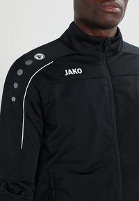 Veste de sport noire en tissu lisse, avec un col haut, des accents blancs et des motifs circulaires sur l'épaule. Logo "JAKO" affiché.