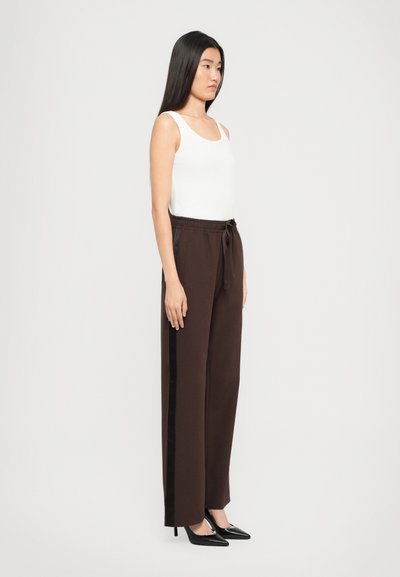 Femme se tenant de profil, portant un top sans manches blanc, un pantalon ample brun avec des rayures sur les côtés, et des escarpins noirs à bout pointu, le tout sur un fond uni.