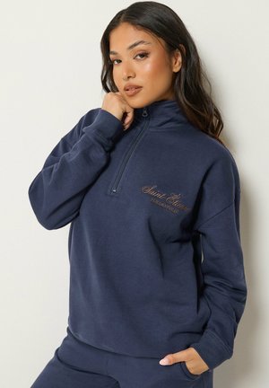 Vrouw met lang donker haar die een marineblauwe sweater met kwart-rits draagt met geborduurde tekst op de borst, poseert met één hand bij haar gezicht.