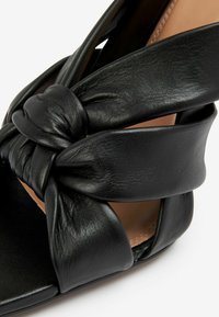 Černé kožené wedge sandály s uzlovým designem, hladkým povrchem a otevřenými boky. Podešev je plochá a minimalistického stylu.