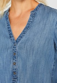 Jasnoniebieska koszula dżinsowa z dekoltem w kształcie litery V, z zapięciem na guziki i widocznymi detalami przeszycia. Materiał wydaje się miękki z lekko teksturowaną powierzchnią.