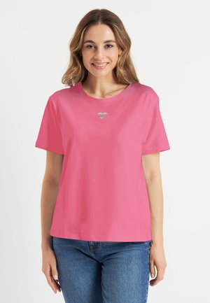 Jonge vrouw met schouderlang haar, die een roze shirt met korte mouwen draagt met een kleine zilveren hartdetail, blauwe spijkerbroek, glimlachend.