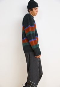 Pleteni pulover v temno zeleni barvi z oranžnimi, teal in modrimi horizontalnimi črtami; mehak material in rebrasti manšeti. Nosijo ga s črnimi hlačami.