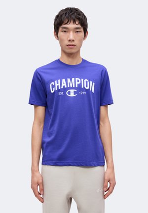 Junger Mann trägt ein lilafarbenes Champion-T-Shirt mit weißem Logo-Schriftzug und hellbeigen Hosen, steht vor einem einfarbigen hellen Hintergrund.