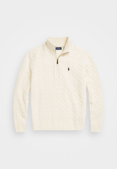 Cream-colored cable-knit sweater with a high collar, featuring a half zipper and a small navy logo on the left side.
