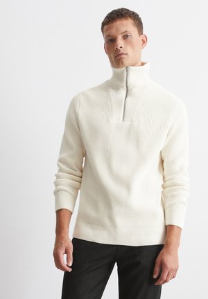 Cream-colored knitted sweater with a high collar, half-zip design, ribbed texture, and fitted sleeves, paired with dark pinstriped trousers.