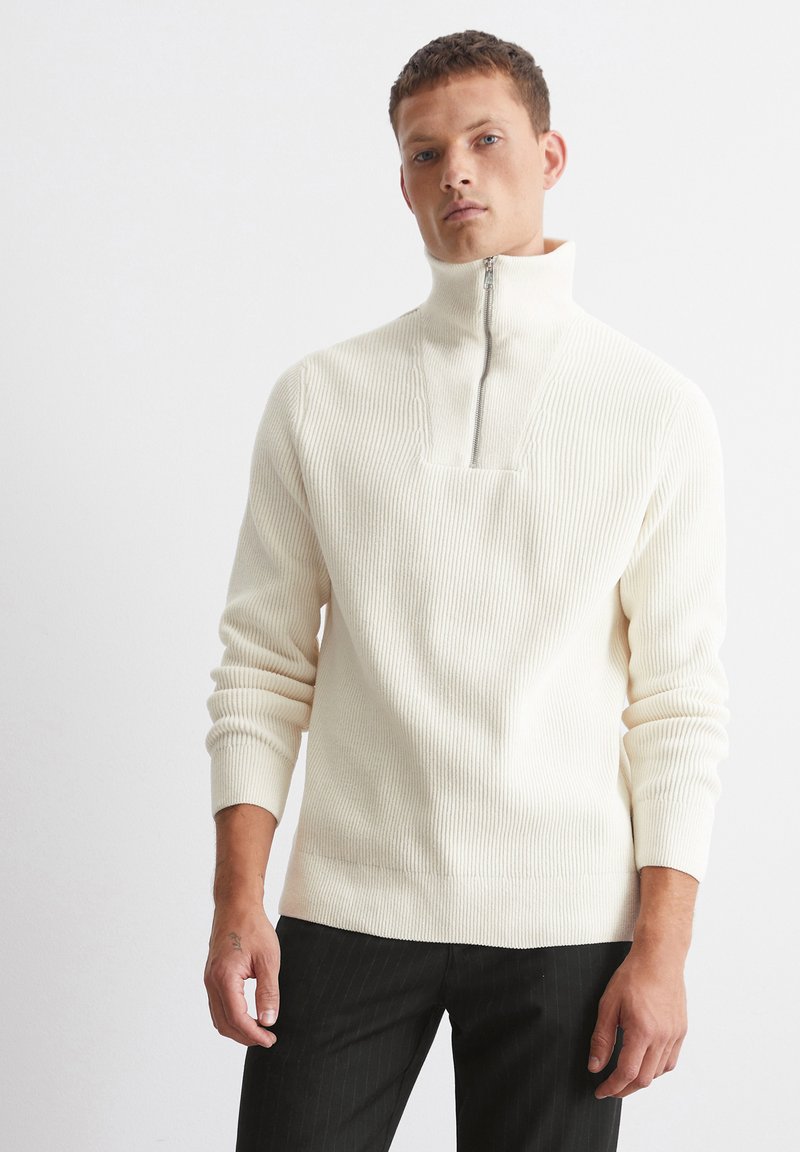 Cremefarbenes gestricktes Sweatshirt mit hohem Kragen, halbem Reißverschluss, gerippter Textur und eng anliegenden Ärmeln, kombiniert mit dunkel gestreiften Hosen.