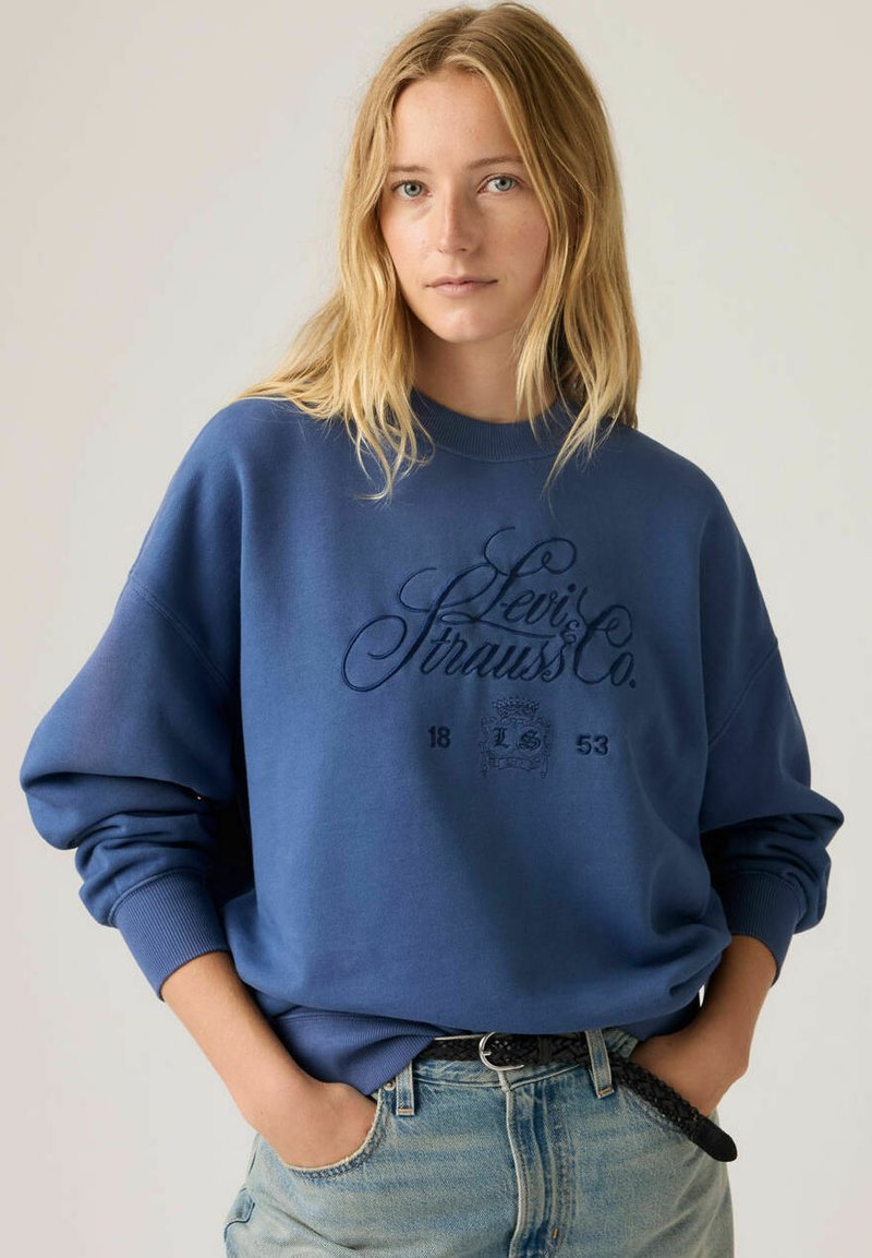 Jeune femme aux cheveux blonds portant un sweat-shirt Levi's bleu et un jean bleu clair, les mains dans les poches, arrière-plan neutre.