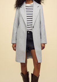 Gray wool coat with a tailored lapel and single button closure, paired with a striped shirt and black distressed shorts. Brown cowboy boots.