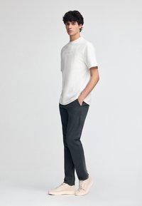 Vit kortärmad t-shirt med präglad text, i kombination med mörkgrå byxor och ljusbeiga sneakers, vilket skapar en avslappnad outfit.