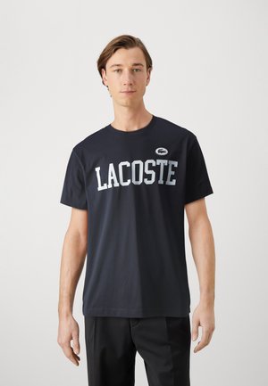 Camiseta de algodón azul marino con el logo blanco de "LACOSTE" en el pecho y un pequeño parche del logo en el hombro. Diseño de manga corta y cuello redondo.
