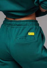 Pantalones de chándal verdes con cintura elástica, bolsillo lateral y una pequeña etiqueta amarilla "LEVEL UP" en la parte trasera. Tejido suave y texturizado.