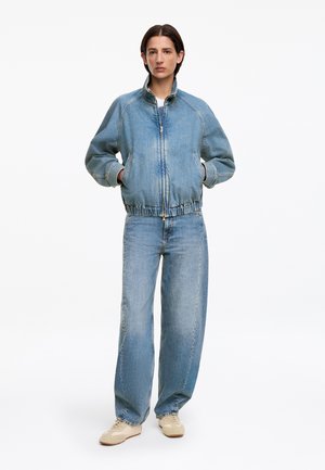 Femme portant une veste en jean claire zippée et un jean large assorti, debout les mains dans les poches sur fond blanc.