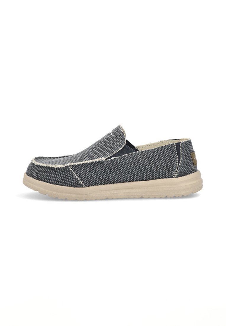 Zapato slip-on de lona en azul marino con superficie texturizada, suela de goma beige en contraste y detalles de costura visibles a lo largo de los bordes.