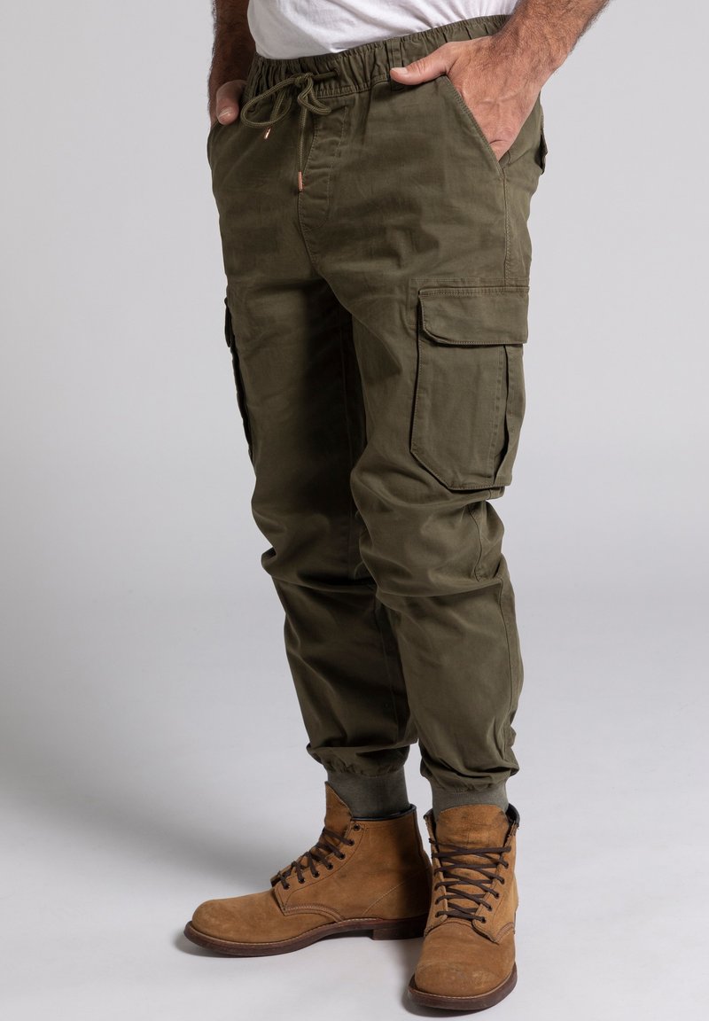 Olivengrüne Cargo-Hose mit elastischem Bund, Kordelzug und seitlichen Taschen. Kombiniert mit braunen Schnürstiefeln.