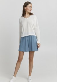 Witte, gestructureerde longsleeve top met een v-hals en een blauwe, loszittende short. Beide kledingstukken hebben een casual pasvorm, gecombineerd met witte sneakers.