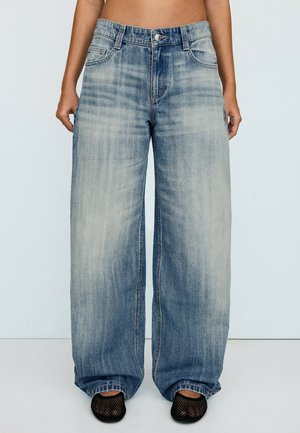 Person iført højtaljede, vidde, falmede blå jeans og sorte mesh slip-on sko, stående mod en ensfarvet, lys baggrund.