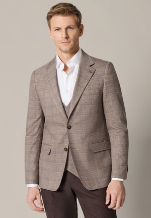 Mężczyzna ubrany w beżową kratkowaną marynarkę na białej koszuli i ciemnobrązowe spodnie na jednolitym beżowym tle.