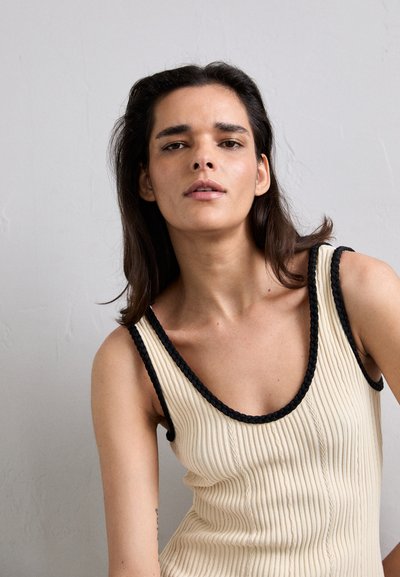 Top de tirantes acanalado crema con ribete trenzado negro a lo largo del escote y las sisas. La tela parece texturizada con líneas verticales y pequeños detalles de botones.