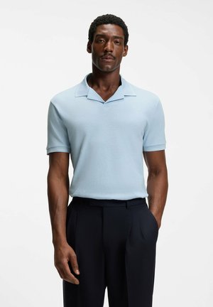 Mies seisoo käsi taskussa, yllään vaaleansininen lyhythihainen kauluspaita ja mustat housut valkoista taustaa vasten.