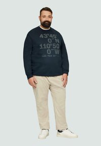 Sweatshirt marine avec des coordonnées imprimées en gris clair, associé à un pantalon beige clair et des baskets blanches. Design simple et décontracté.