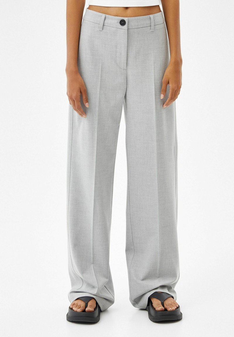 Personne portant un pantalon large taille haute gris clair et des sandales à plateforme noires avec les orteils visibles, sur un fond blanc uni.