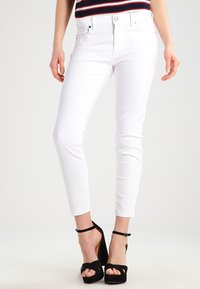 Jeans blancos con un corte ajustado, diseño clásico de cinco bolsillos y un ligero estiramiento. Combinados con sandalias negras de tacón alto.