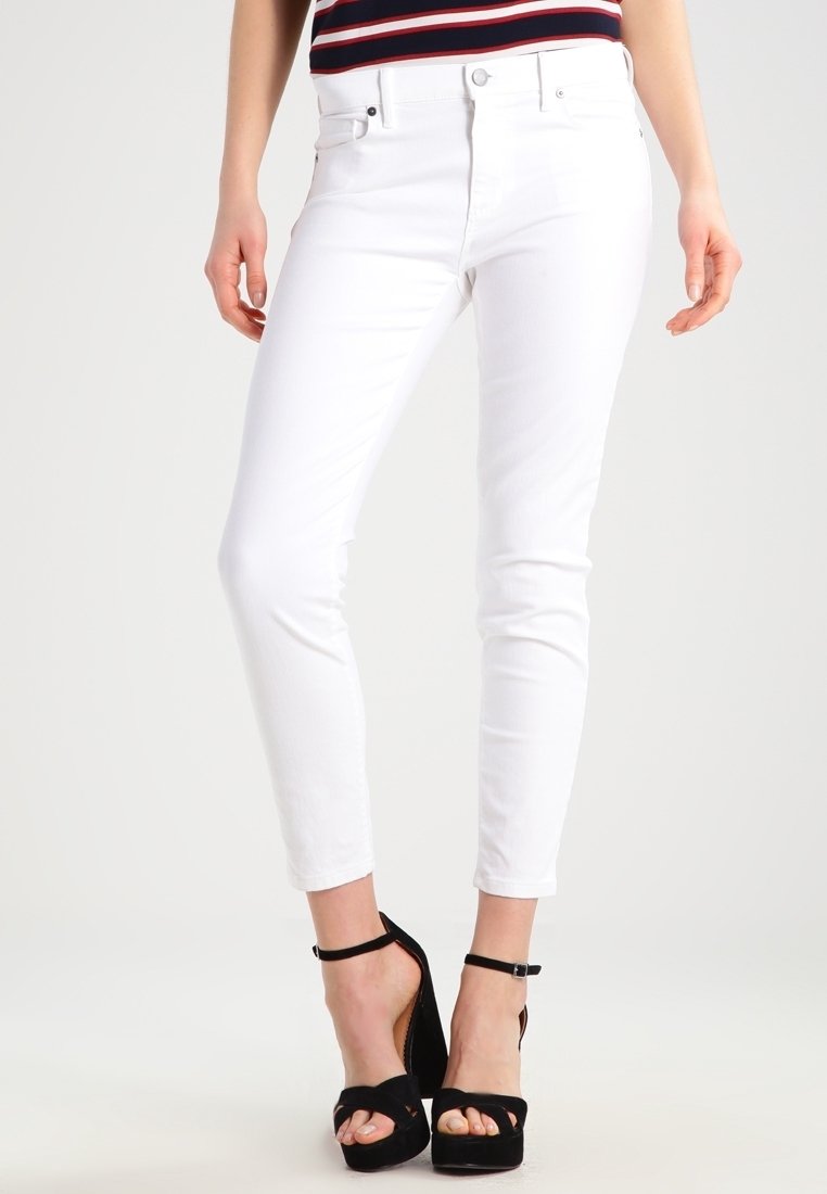 Jeans blancos con un corte ajustado, diseño clásico de cinco bolsillos y un ligero estiramiento. Combinados con sandalias negras de tacón alto.
