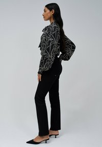 Blusa negra con patrón abstracto en blanco, con cuello volado, combinada con pantalones ajustados negros y tacones de punta negra, vista de lado.