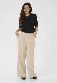 Haut noir à manches courtes associé à un pantalon large beige, présentant une texture lisse, une taille ajustée et des poches avant. Chaussures noires à bout pointu.