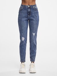 Foranbilde av en person som har på seg blå høytlivs jeans med riper og hvite joggesko mot en ensfarget hvit bakgrunn.