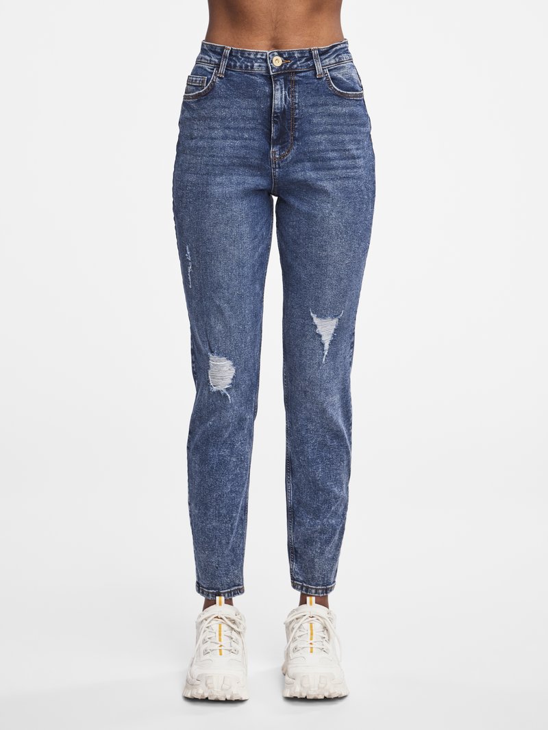 Foranbilde av en person som har på seg blå høytlivs jeans med riper og hvite joggesko mot en ensfarget hvit bakgrunn.