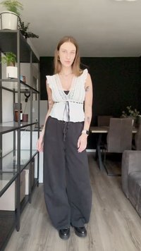 Blusa bianca con rifiniture in pizzo e maniche a volant, legata in vita, abbinata a pantaloni neri a gamba larga e scarpe con zeppa.