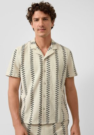 Mężczyzna z kręconymi włosami, ubrany w beżową koszulę na krótki rękaw z czarnymi pionowymi paskami w zygzak, stoi na tle jednolitego jasnego tła.