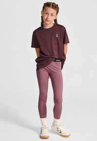 Bordeauxrød t-shirt med korte ærmer og et lille emblem, kombineret med lyse lilla leggings og hvide sneakers med beige detaljer.