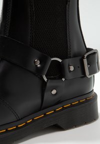 Botte en cuir noir avec des coutures jaunes, panneau latéral élastique et anneau décoratif en métal avec sangle à boucle.