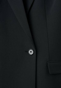 Crni blejzer od tkanine s glatkom teksturom, s blagim sjajem. Zatvaranje na jedno okruglo dugme s dizajnom od četiri rupe i detalj malog džepa.