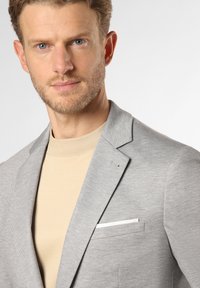 Hellgrauer strukturierter Blazer mit Revers und schmaler Passform, kombiniert mit einem beigen Hochkragenhemd. Verfügt über eine Brusttasche mit weißem Akzent.