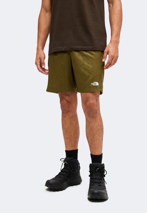 Mann trägt olivgrüne The North Face Shorts, schwarze Socken und schwarze Wanderschuhe, steht vor einem schlichten hellen Hintergrund.