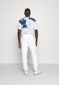Man i vita joggers, vita sneakers med beige detaljer, och en blå och vit tie-dye T-shirt står mot en neutral vägg med ryggen vänd.